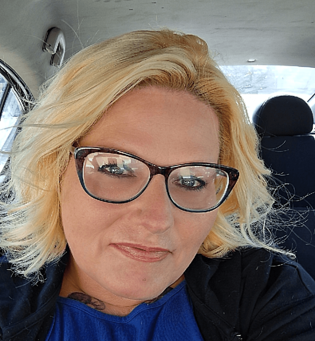 Woman with blonde hair and glasses sitting in a car, smiling, wearing a blue top and black jacket.