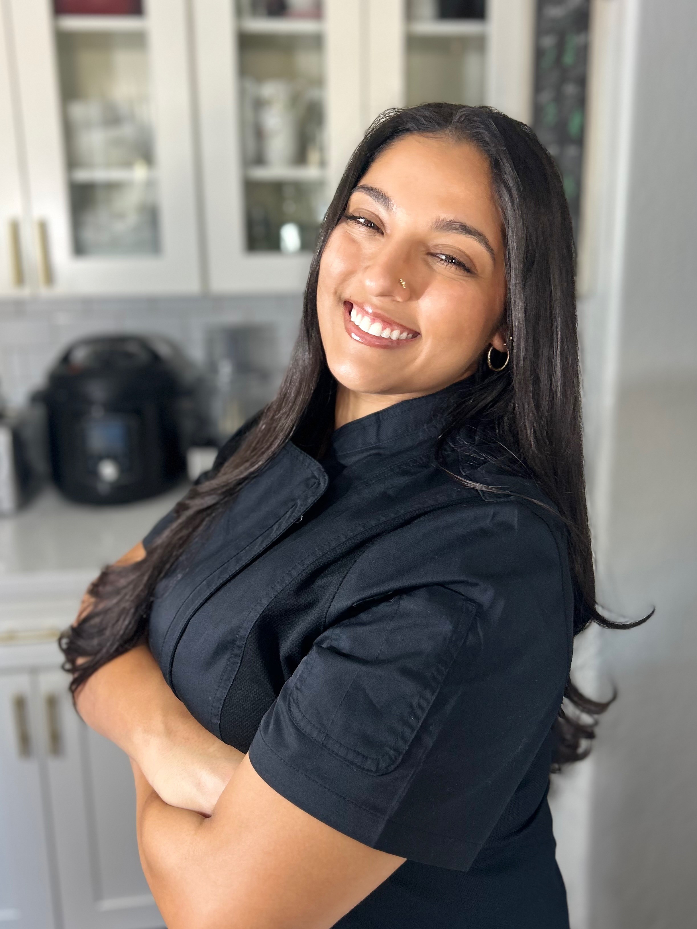 Young woman with long dark hair smiling in a kitchen, arms crossed, wearing a black top.