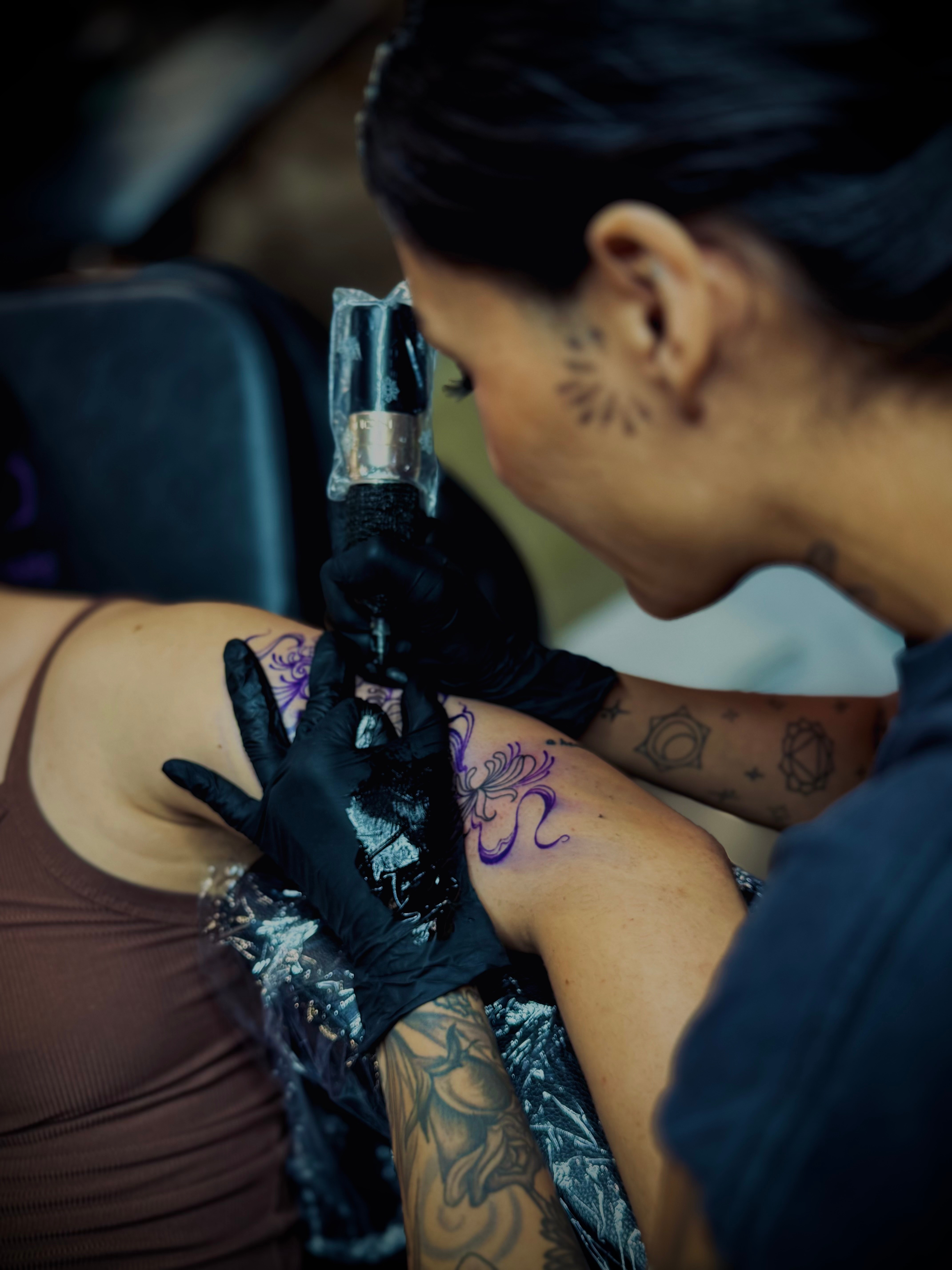 Tattoo artist with black gloves tattooing a purple design on a person's arm, woman with dark hair and face tattoo, focused.