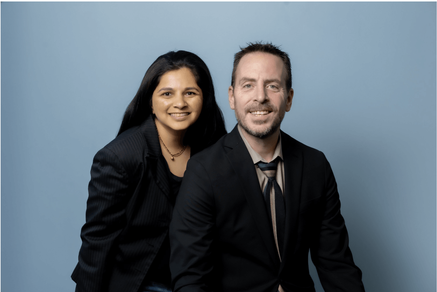 Two people, a woman and a man, pose together against a light blue background, smiling at the camera.