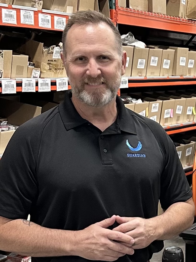 Man with short hair and beard wearing a black polo shirt with a logo, standing in a warehouse with shelves of boxes.