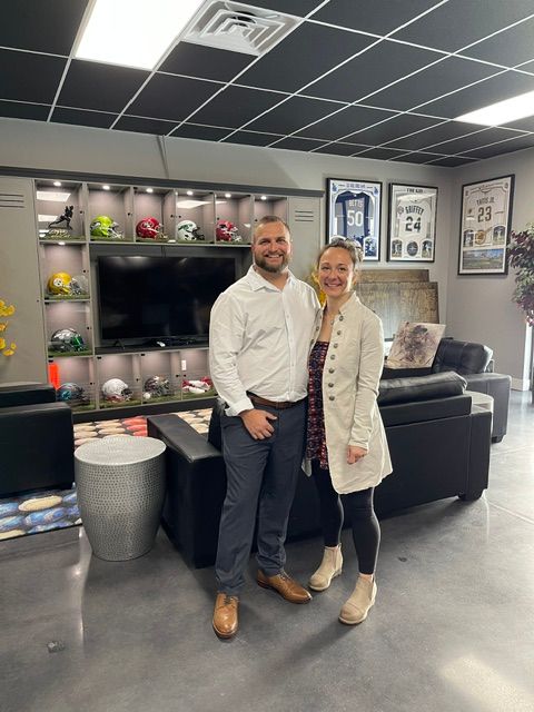 Two men standing in a modern office lounge area with shelves, a TV, and seating, one in a white shirt and the other in a beige coat.