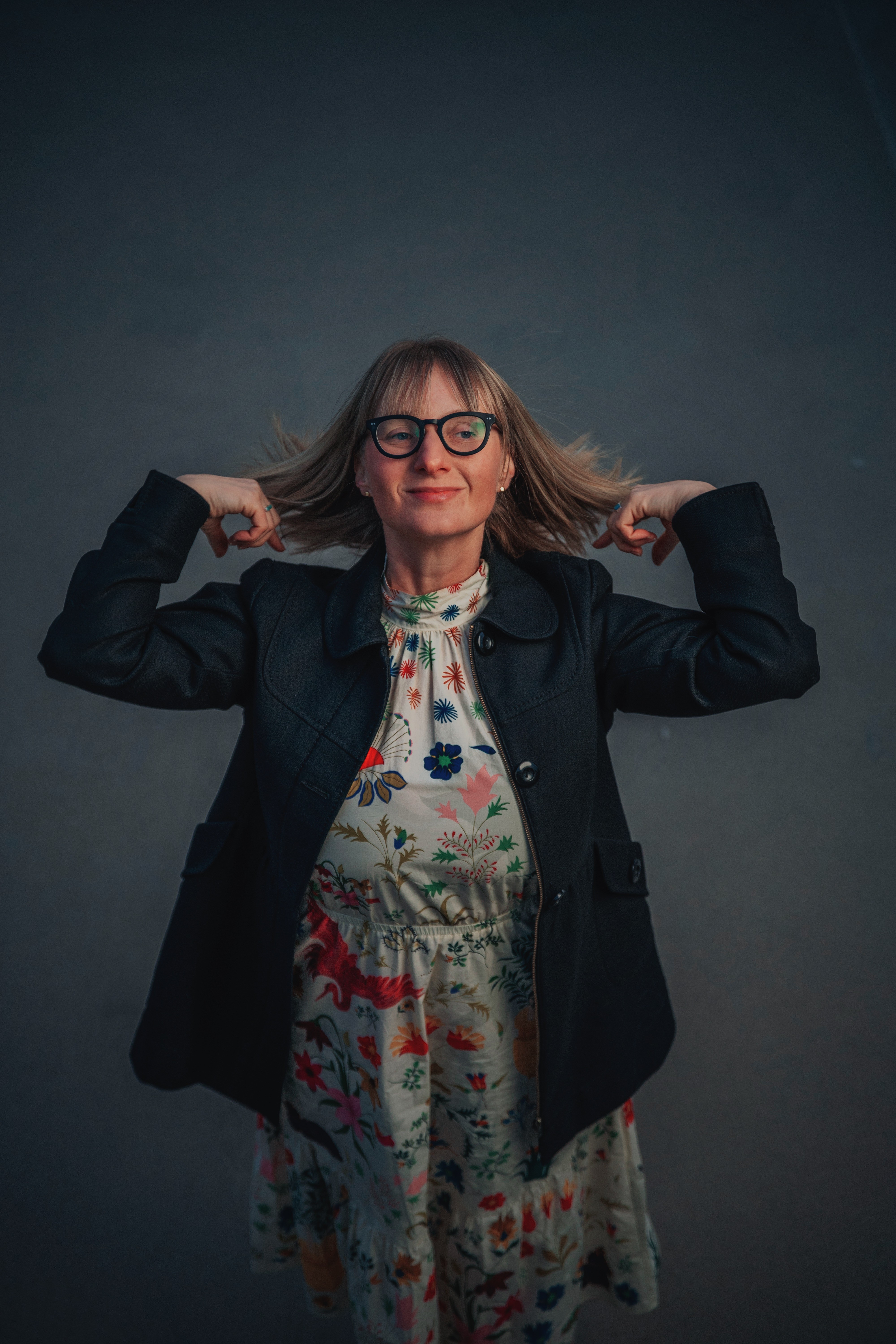 Woman with glasses and shoulder-length hair, wearing a floral dress and dark jacket, poses with hands behind her head against a dark background.