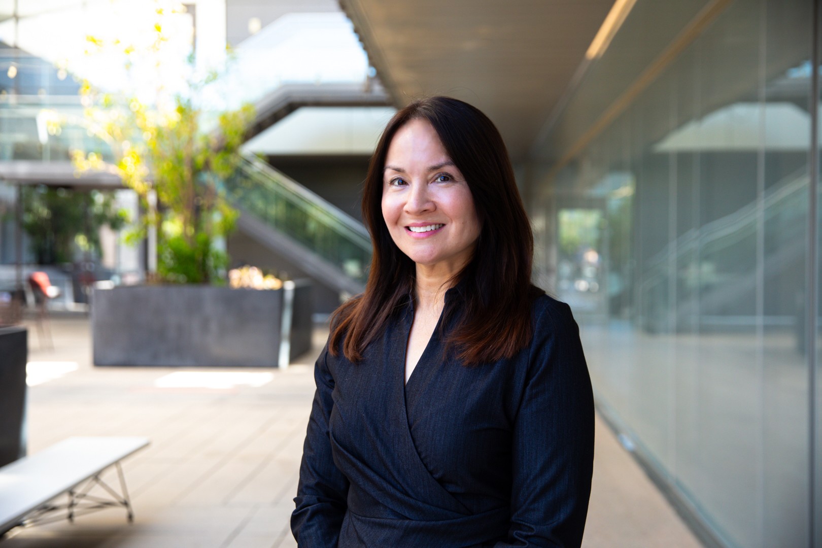 Woman with dark hair smiling, wearing a dark blazer, standing outdoors near a modern building with glass walls.