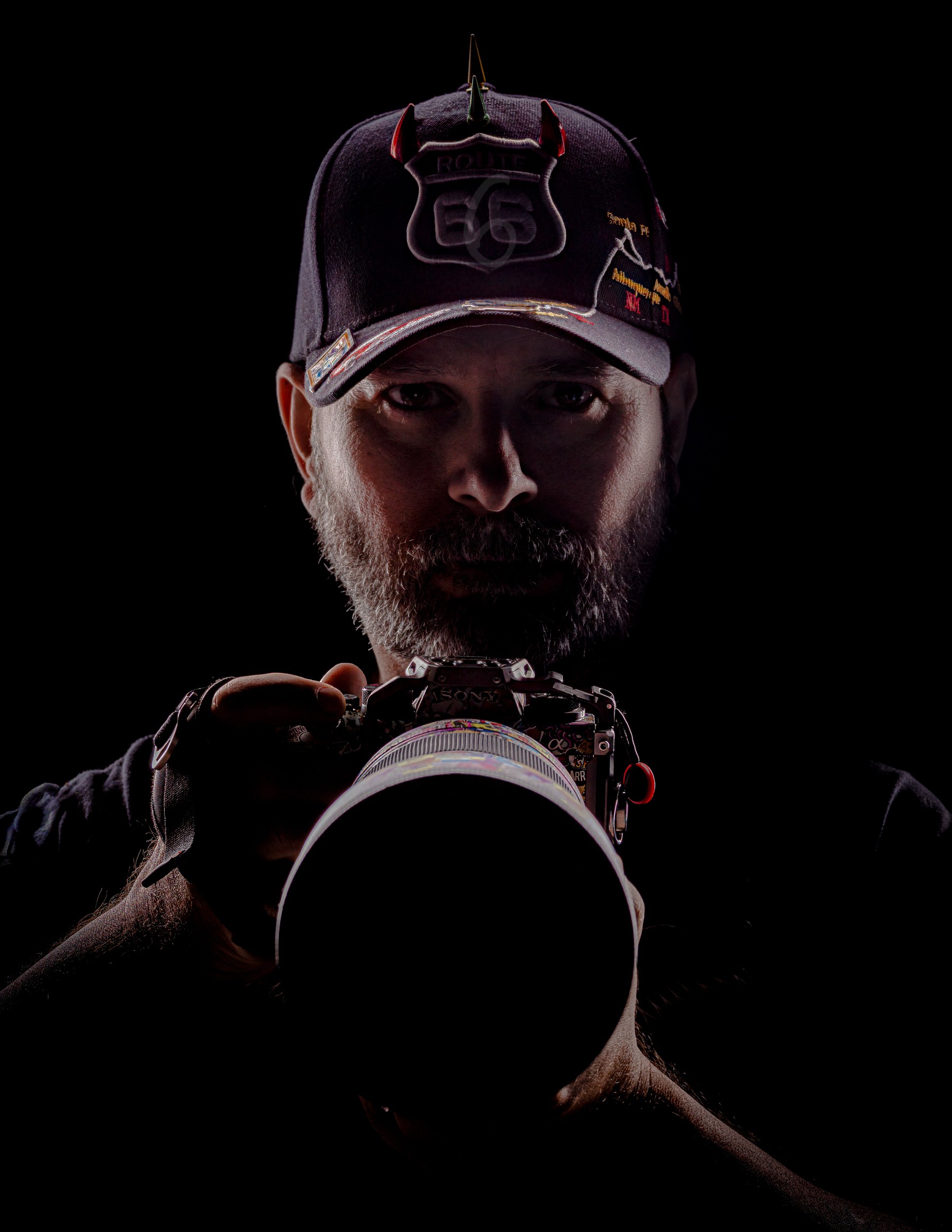 Person wearing a cap, holding a camera, with a dark background, face partially illuminated.