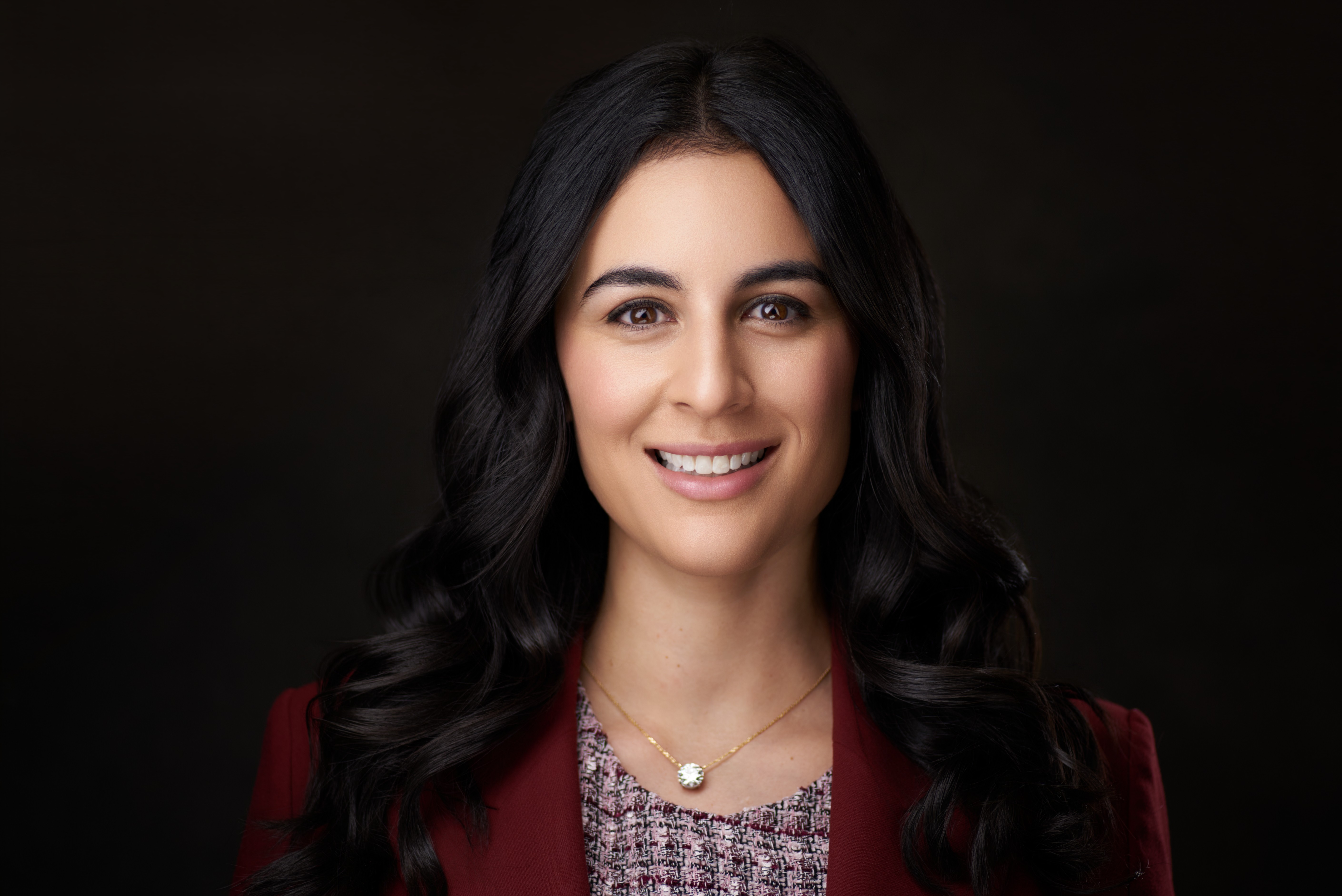 Portrait of a smiling woman with long dark hair, wearing a red blazer and a patterned top, against a dark background.