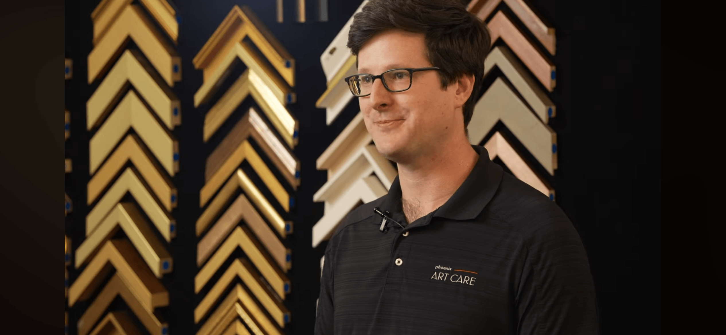 Man with glasses and dark hair wearing a black polo shirt stands in front of a display of picture frame samples.