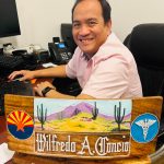 Man smiling at desk with computer and phone, holding a wooden plaque with desert scene and symbols, in an office.