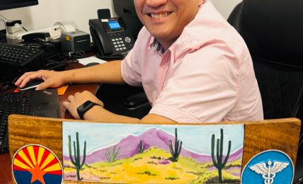 Man smiling at desk with computer and phone, holding a wooden plaque with desert scene and symbols, in an office.
