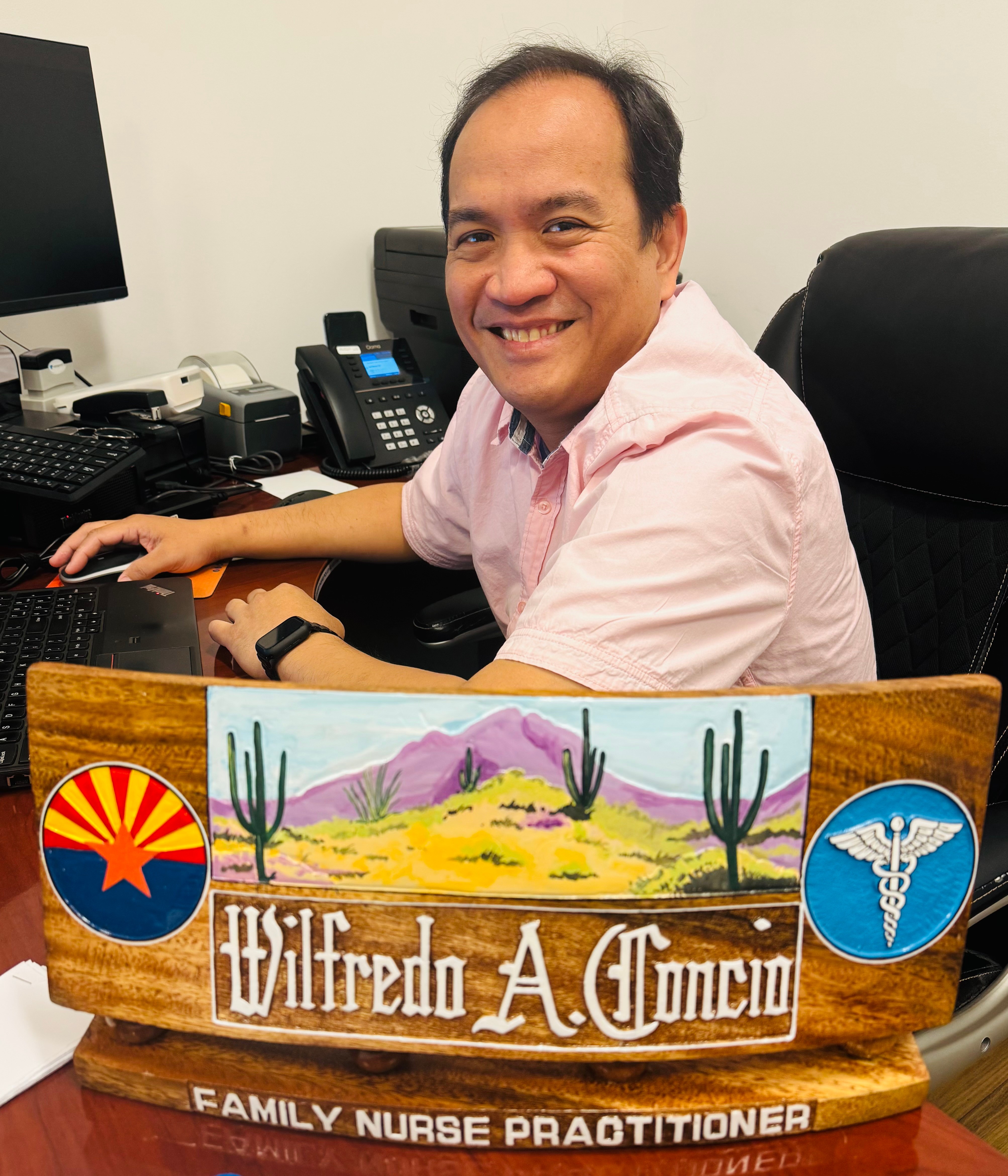Man smiling at desk with computer and phone, holding a wooden plaque with desert scene and symbols, in an office.