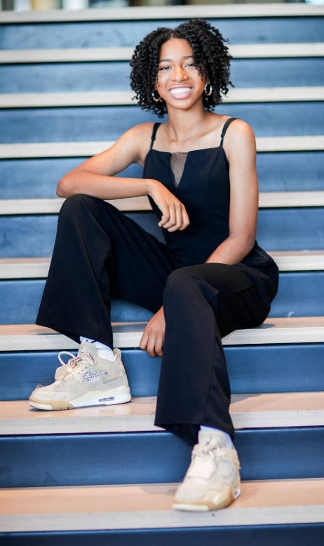 Young woman with curly hair sitting on stairs, smiling, wearing a black jumpsuit and sneakers.