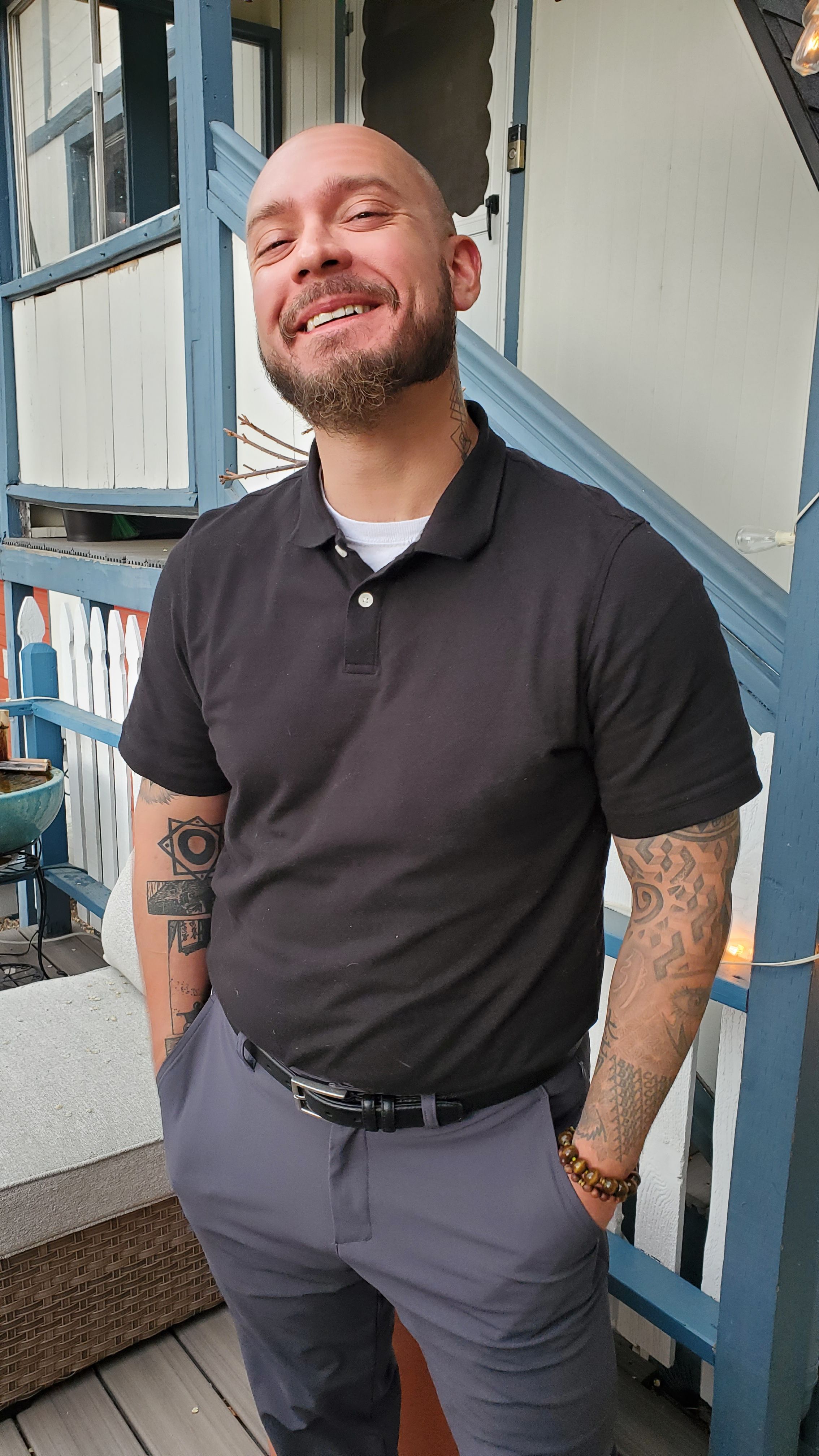 Man smiling, wearing black shirt and gray pants, standing outdoors near a blue structure.