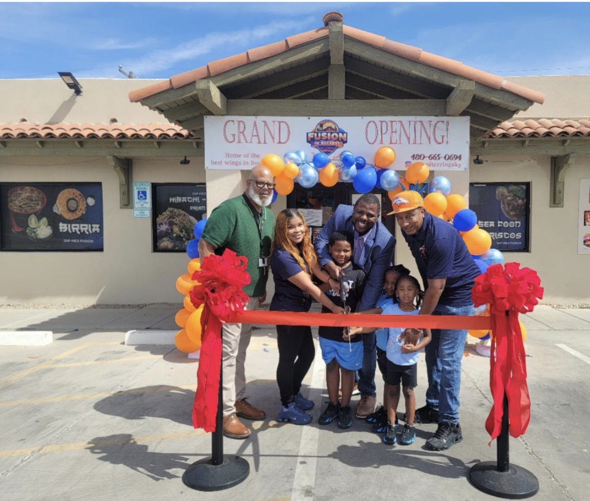 Group of people at grand opening with balloons and ribbon outside a building, smiling and holding scissors, children in front.