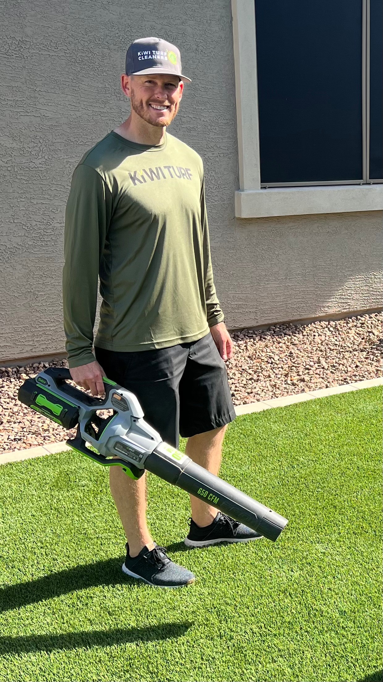 Person standing outdoors on grass holding a leaf blower, wearing a cap, long-sleeve shirt, and shorts.