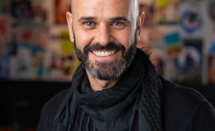 Bald man with beard smiling, wearing a black shirt and scarf, in front of a colorful collage background.