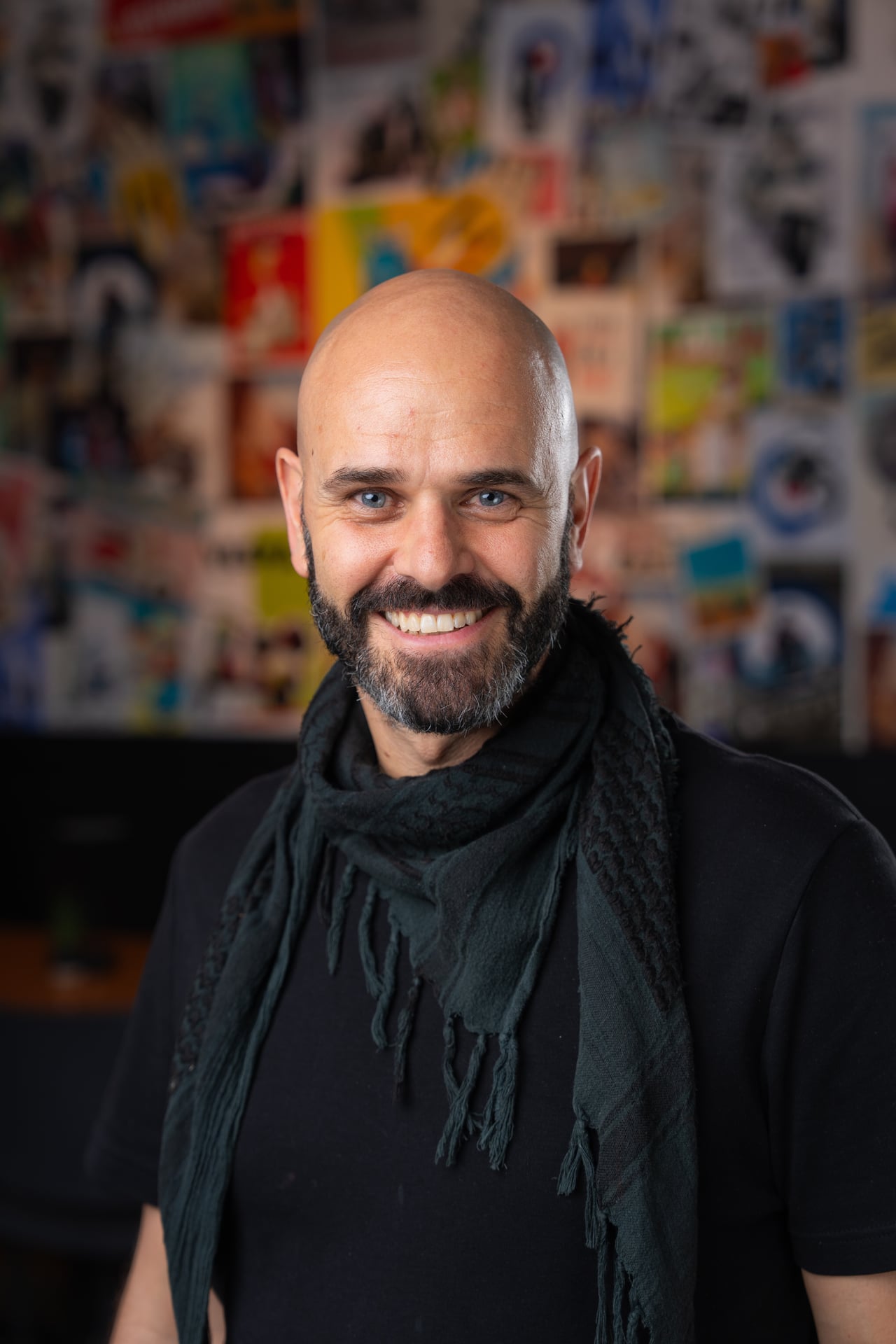 Bald man with beard smiling, wearing a black shirt and scarf, in front of a colorful collage background.