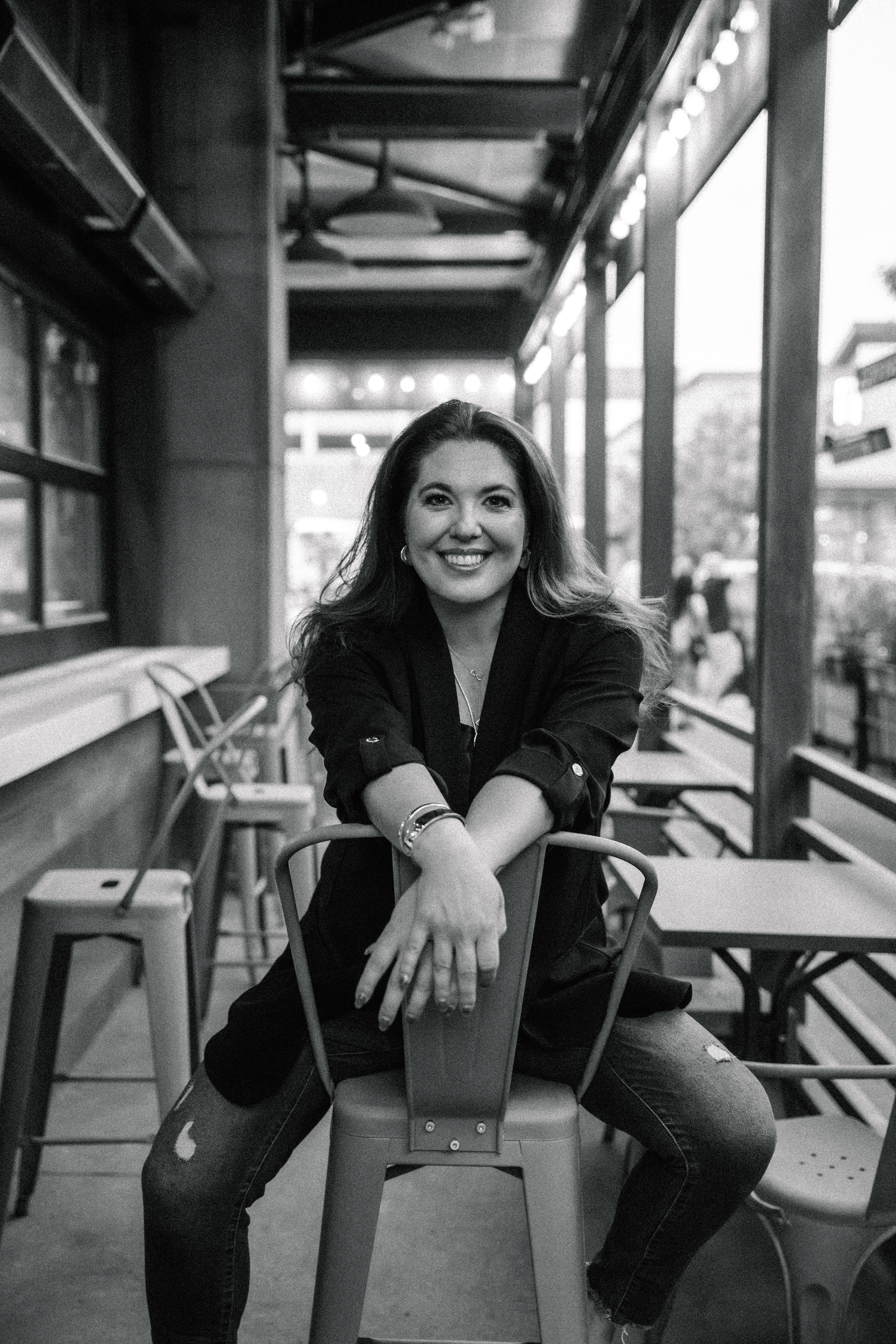 Woman sitting on a chair in a cafe, smiling, with large windows and outdoor seating visible behind her.