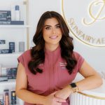 Woman in pink uniform smiling, standing in skincare clinic with shelves of products behind her.