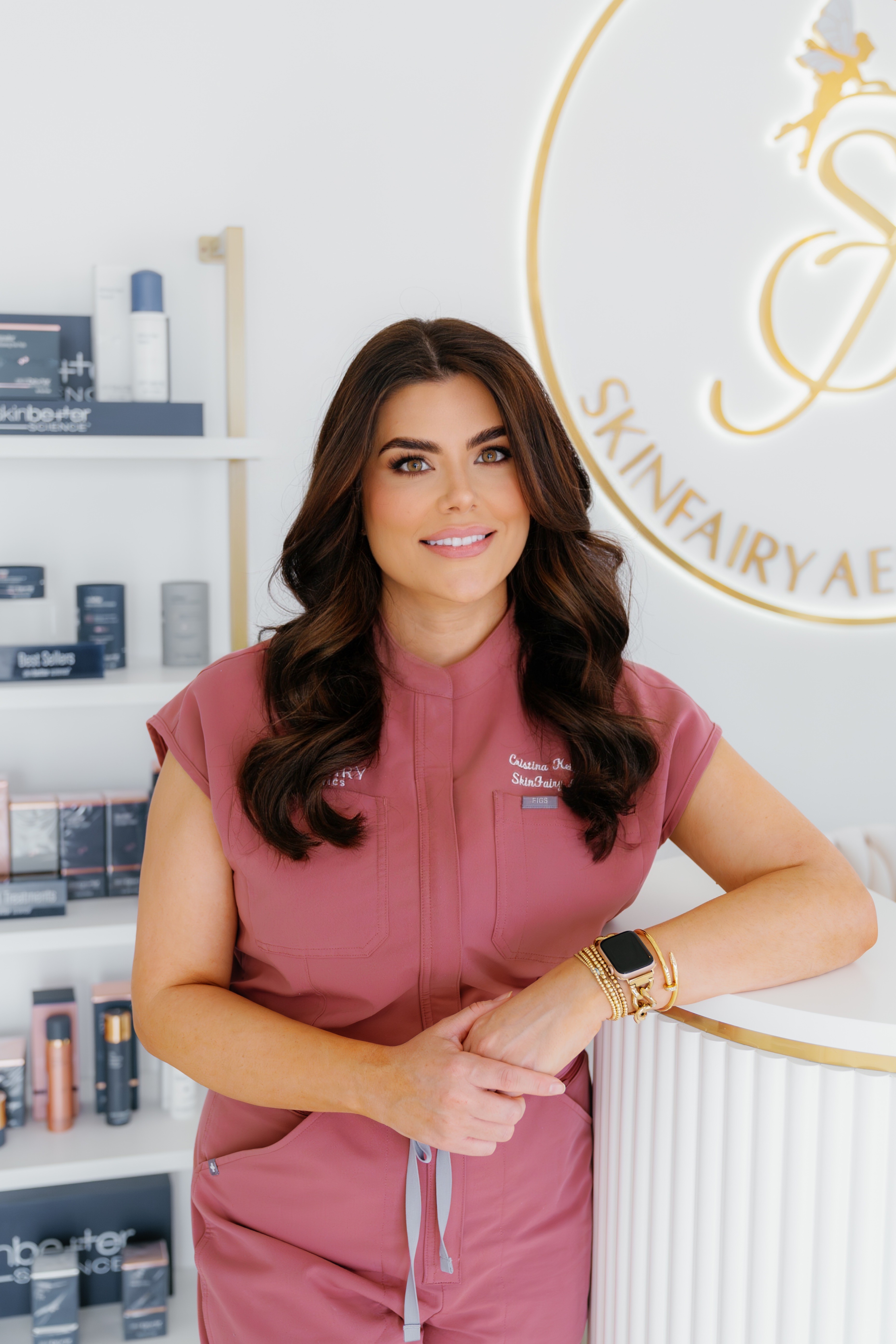 Woman in pink uniform smiling, standing in skincare clinic with shelves of products behind her.