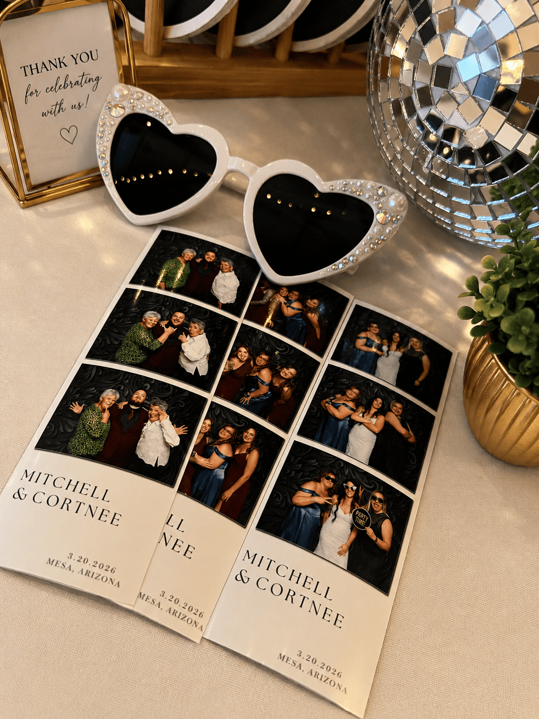 Two photo booth strips with people posing, heart-shaped sunglasses, a thank you card, a disco ball, and a potted plant on a table.