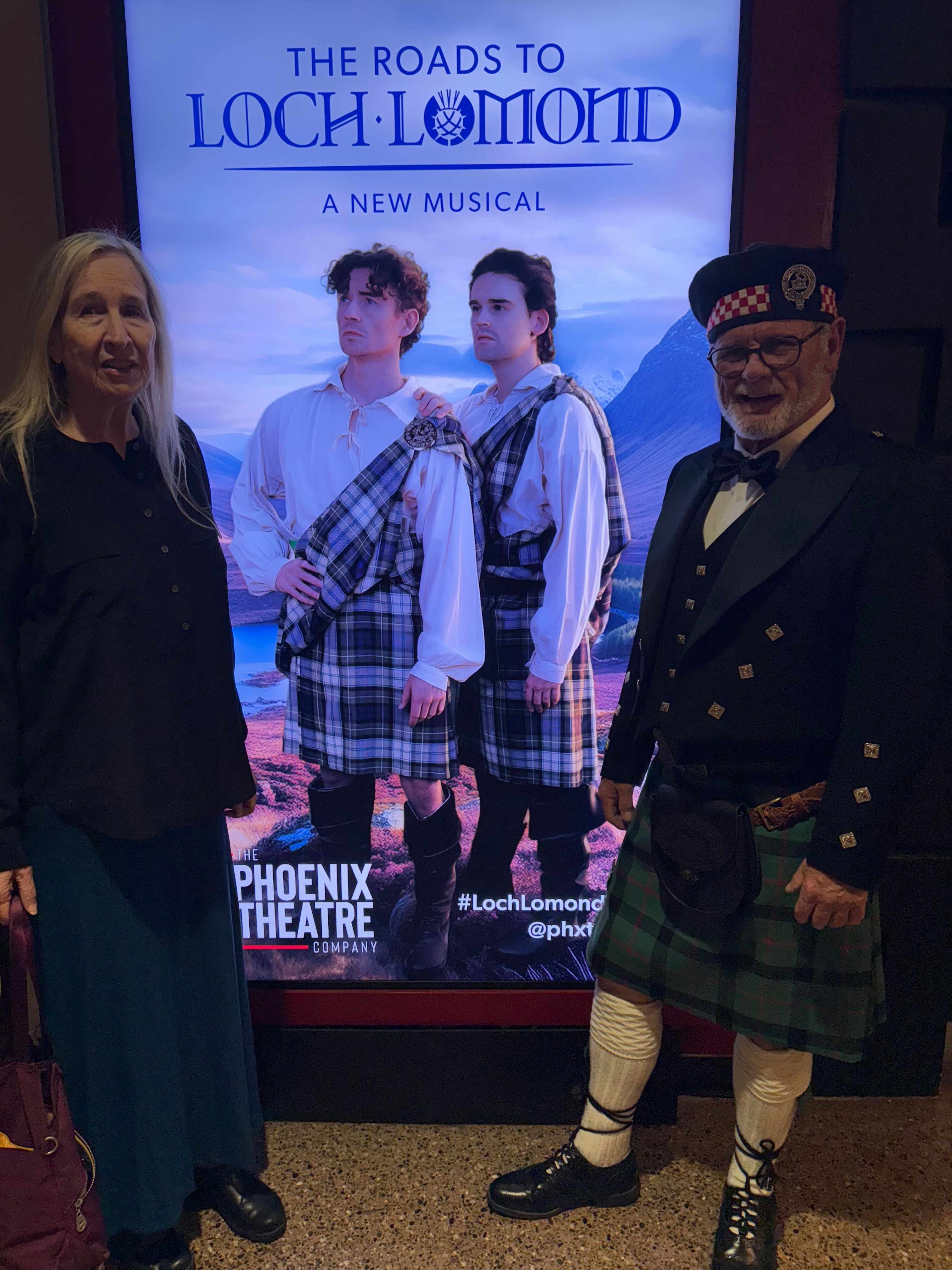 Two people standing in front of a digital screen displaying a promotional poster for a theatrical production, with one person wearing a kilt and the other in dark clothing.