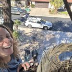 Person smiling outdoors with a view of a street, parked cars, and two people standing on the sidewalk.
