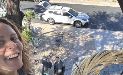 Person smiling outdoors with a view of a street, parked cars, and two people standing on the sidewalk.
