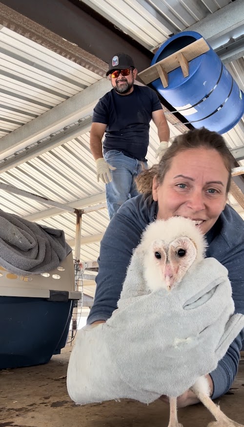 Woman holding a small white puppy with a person standing in the background in an industrial setting.