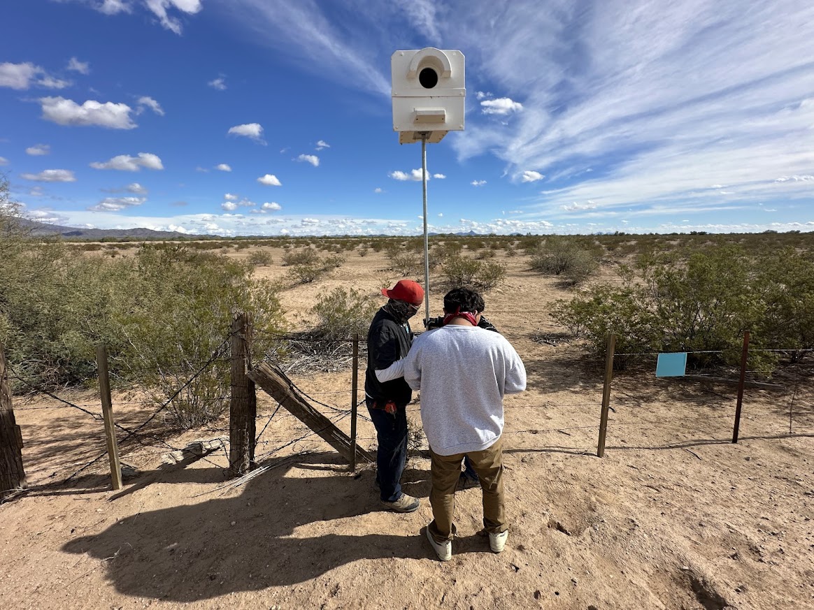 Two people stand outdoors near a fence, looking at a large white device mounted on a pole against a desert landscape with bushes and a blue sky.