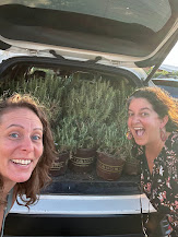 Two women smiling in front of a car with potted plants in the trunk, outdoors on a sunny day.