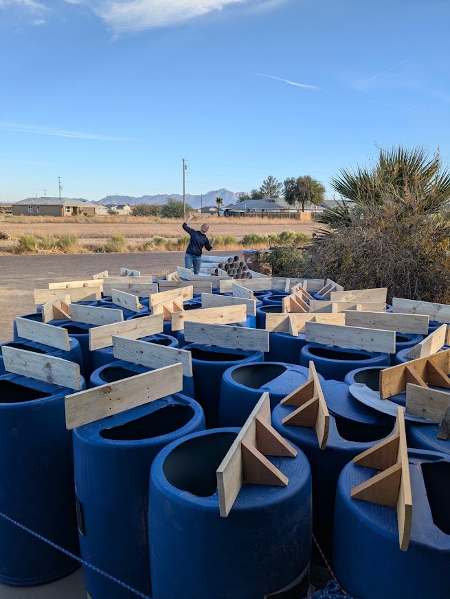 Person working outdoors with large blue pipes and wooden supports, desert landscape, mountains, and clear sky in background.