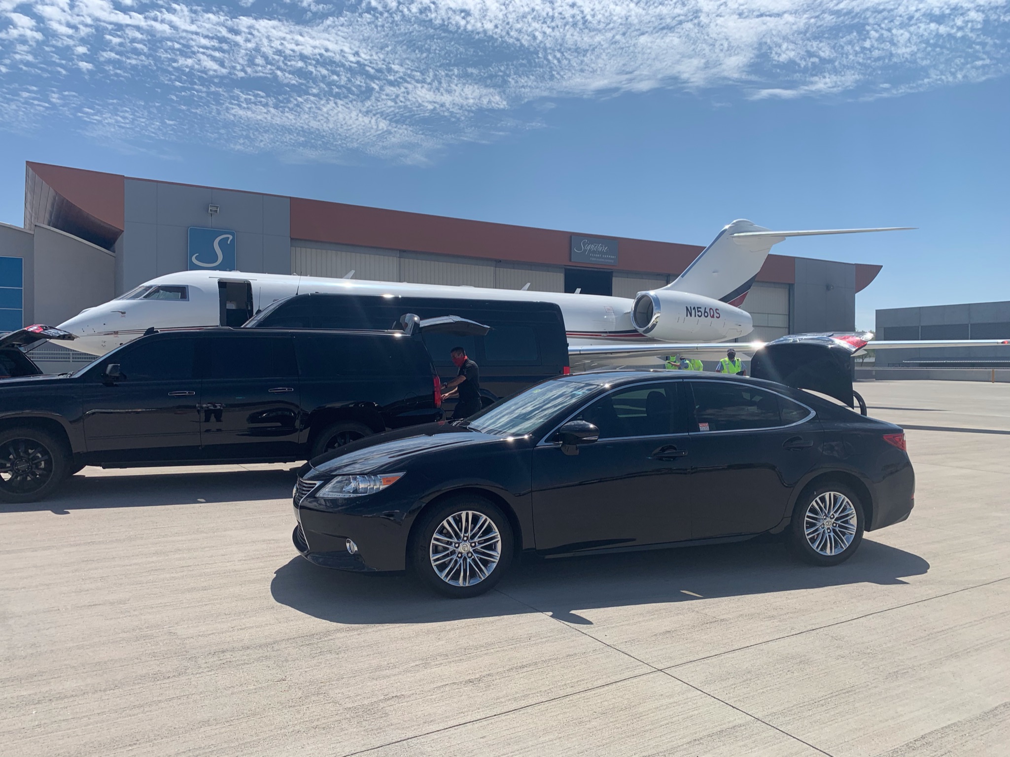 Private jet on tarmac with two black cars parked nearby, airport building in background, clear sky.