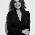 Woman with shoulder-length wavy hair smiling, wearing a black blazer and top, standing against a plain background.