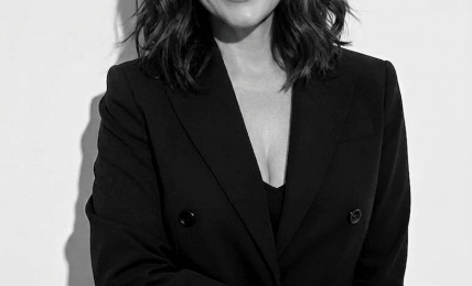 Woman with shoulder-length wavy hair smiling, wearing a black blazer and top, standing against a plain background.