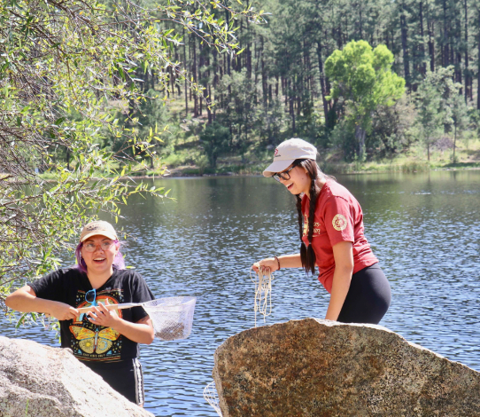 Two girls by a lake, one holding a net and the other pouring water, surrounded by trees and rocks.