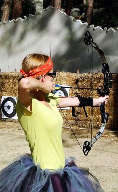 Child with a red bandana practicing archery outdoors, aiming with a bow and arrow, target visible in background.