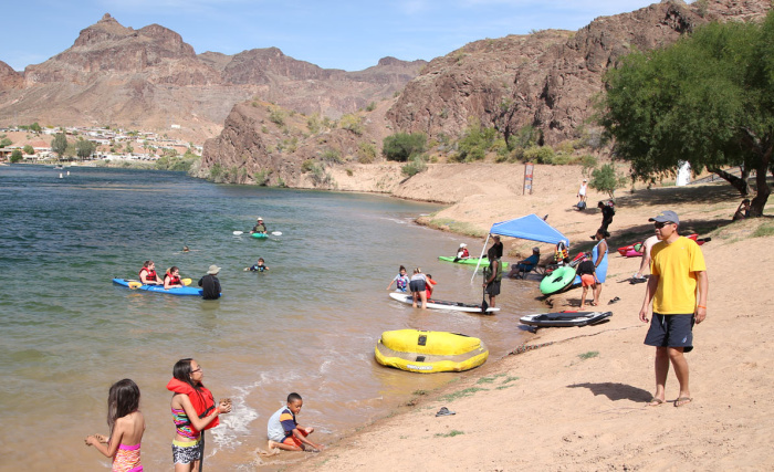 People enjoying a day at a lakeside beach with boats, a yellow float, and mountains in the background.