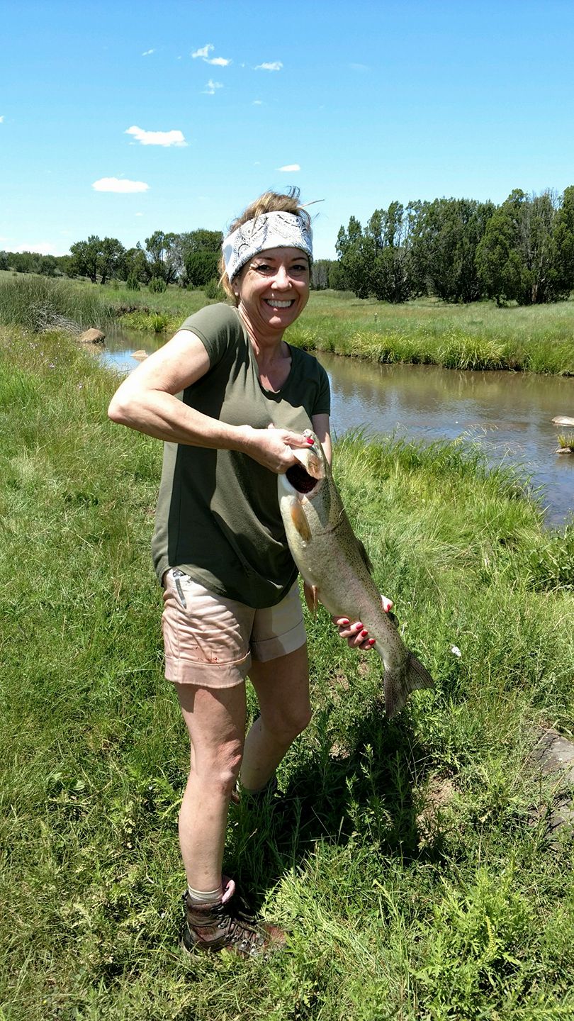 Woman outdoors holding a large fish near a river with green grass and trees, blue sky, smiling, wearing a headband and casual clothes.