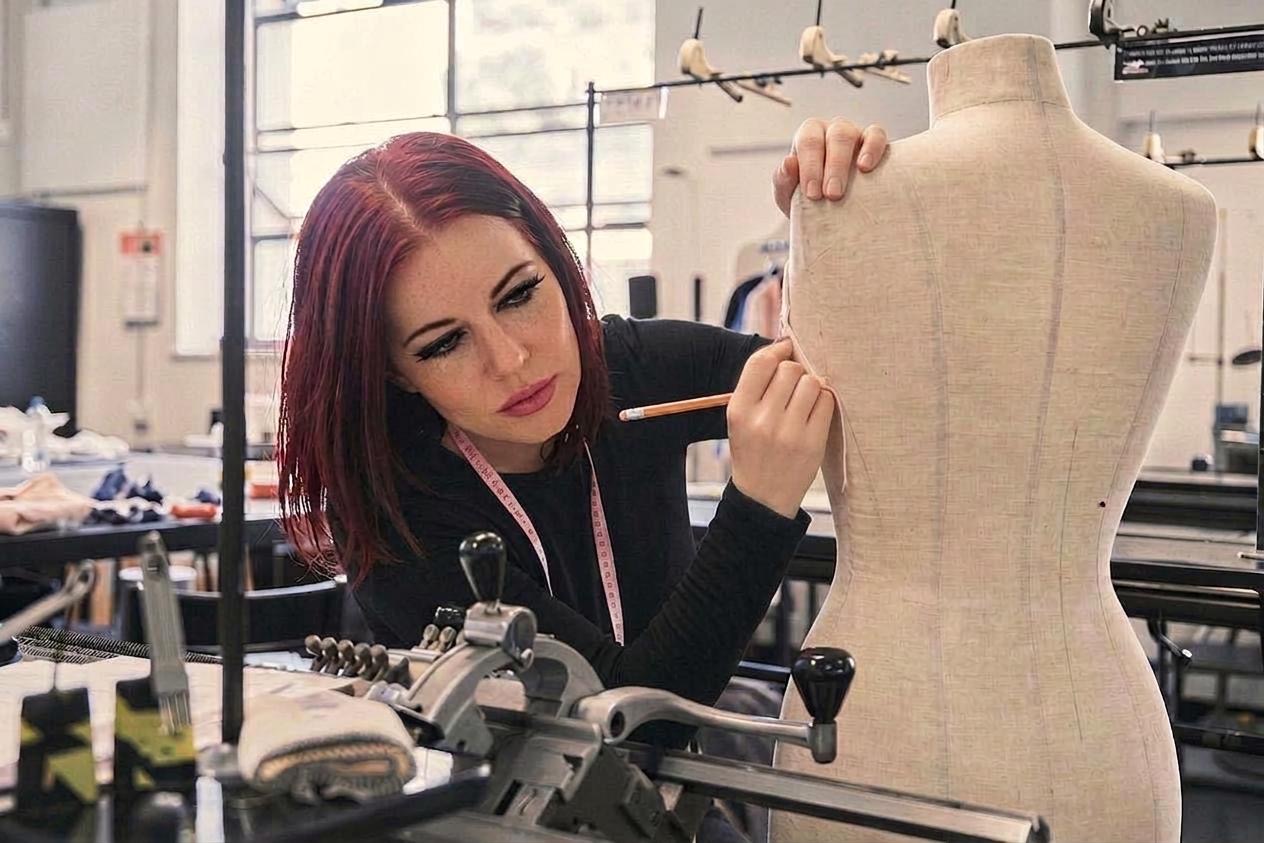 Woman with red hair working on a dress form in a sewing studio, surrounded by sewing tools and fabric.