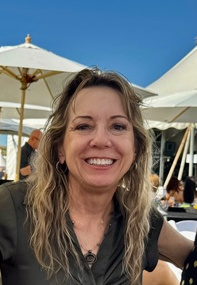 Woman smiling outdoors with umbrellas and people in background, sunny day, blue sky.