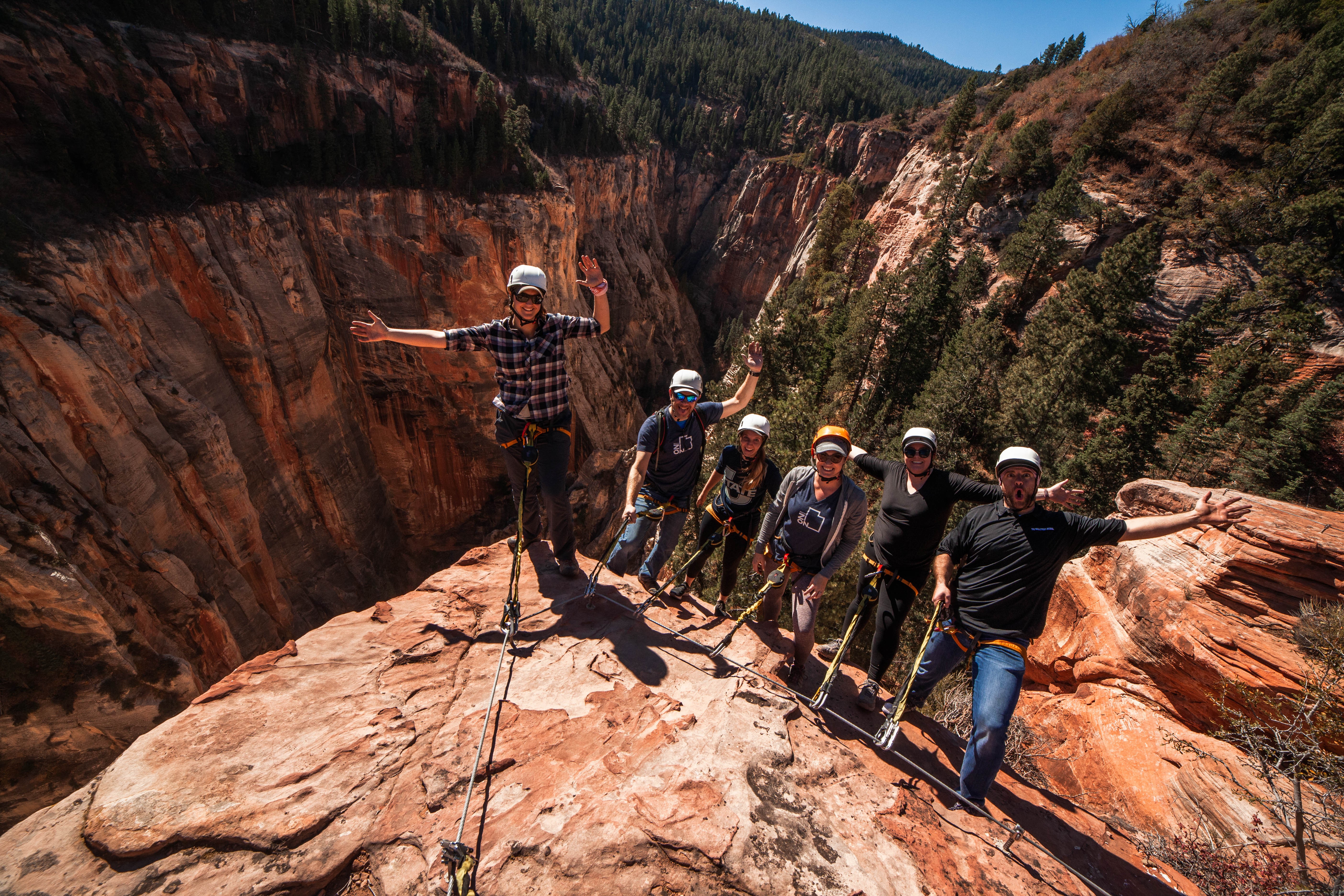 Group of six people hiking on a rocky ledge in a canyon with steep walls and trees, holding trekking poles.