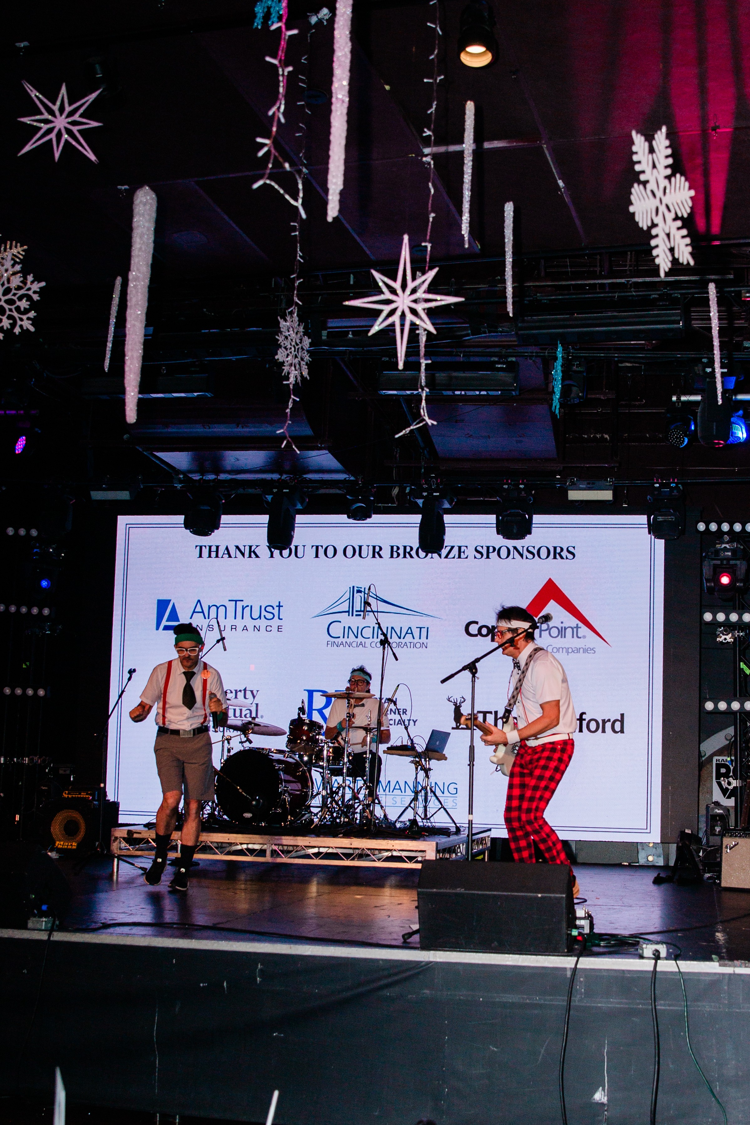 Band performing on stage with holiday decorations hanging above, including stars and snowflakes, in a dark venue with a large screen behind.