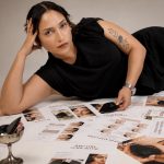 Woman lying on her side on a table covered with magazines, resting her head on her hand, wearing a black top, with tattoos on her arm.