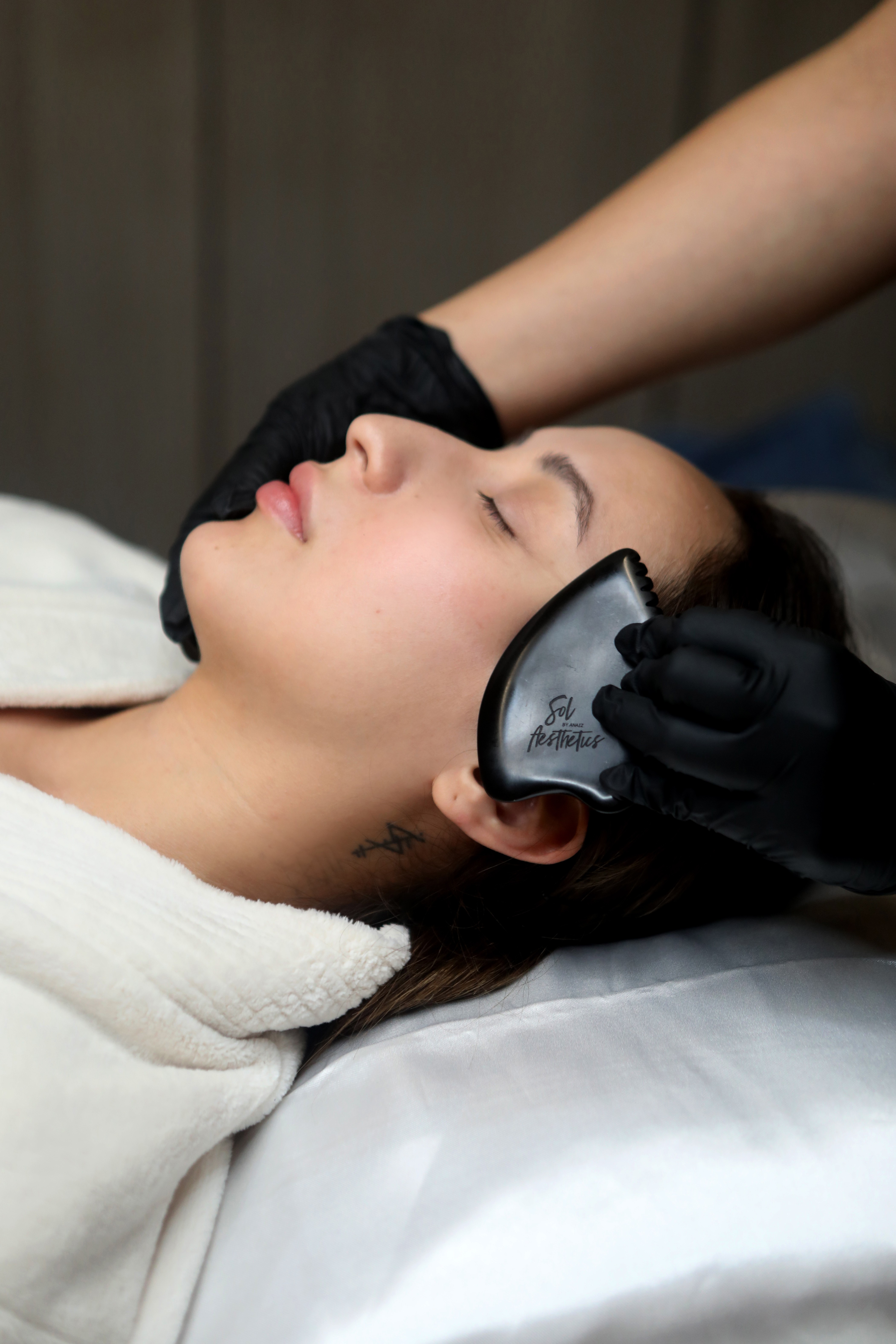 Person receiving a facial treatment with eyes closed, lying down, and a technician applying a mask or cream.