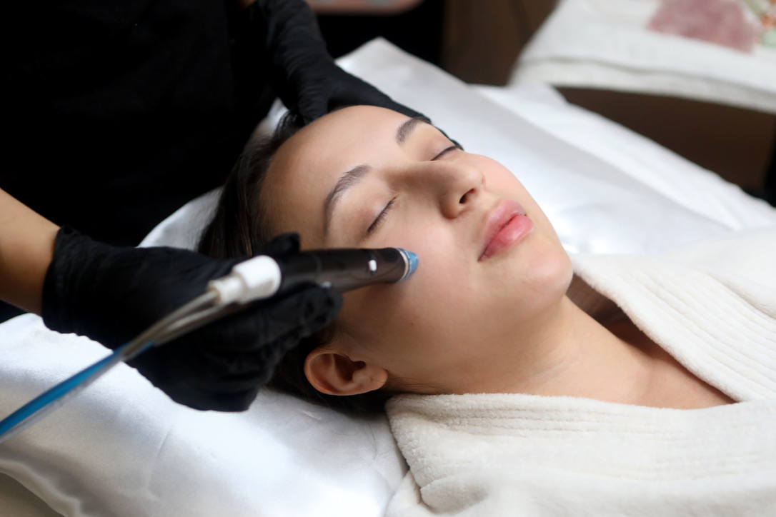 Woman receiving facial treatment with a device on her face, lying on a bed with eyes closed, in a spa or clinic setting.