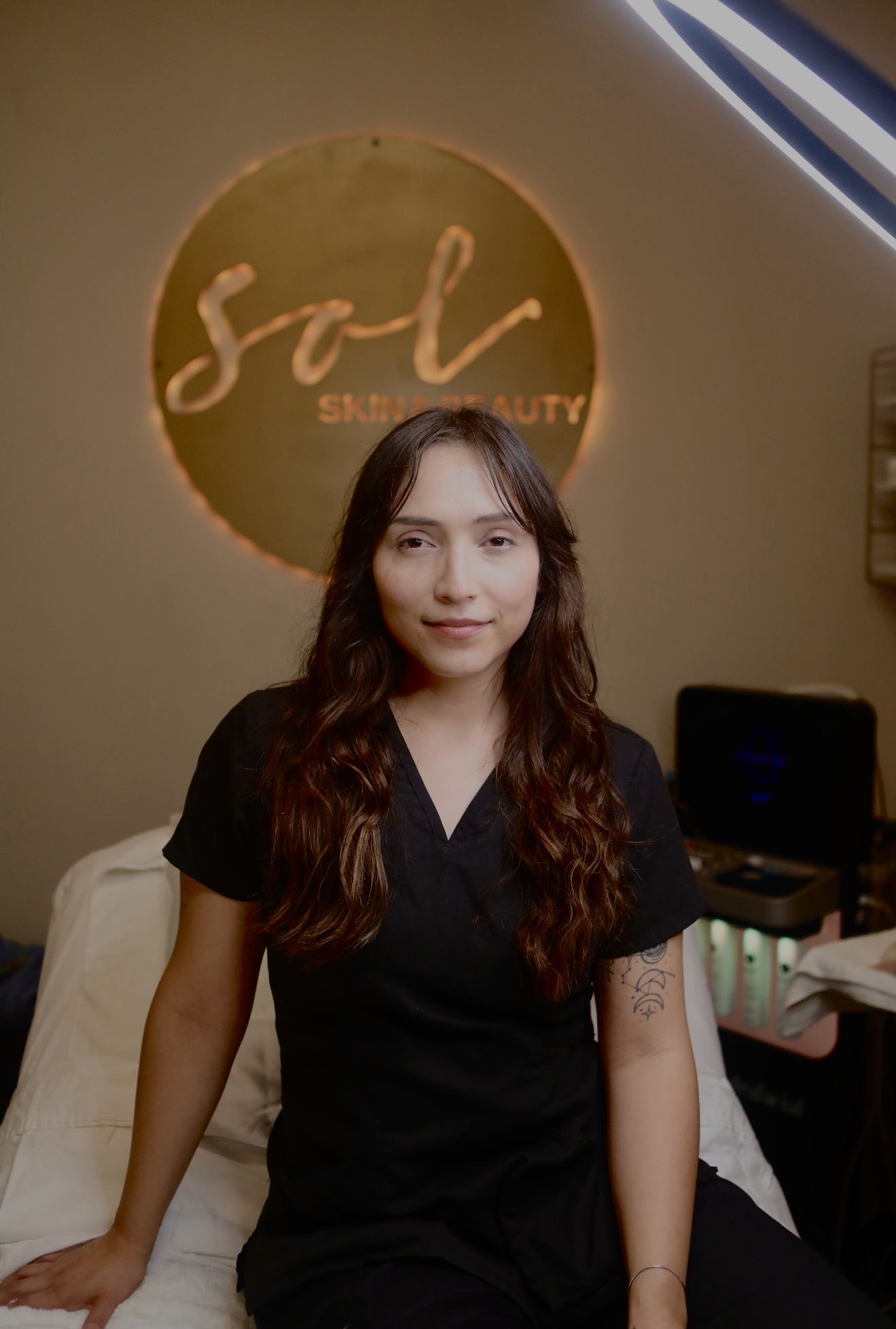 Young woman with long wavy hair in black scrubs standing in front of a wall with a circular logo reading 'sol' and 'BEAUTY'.