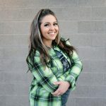 Smiling woman with long hair wearing a green plaid shirt standing against a gray brick wall.