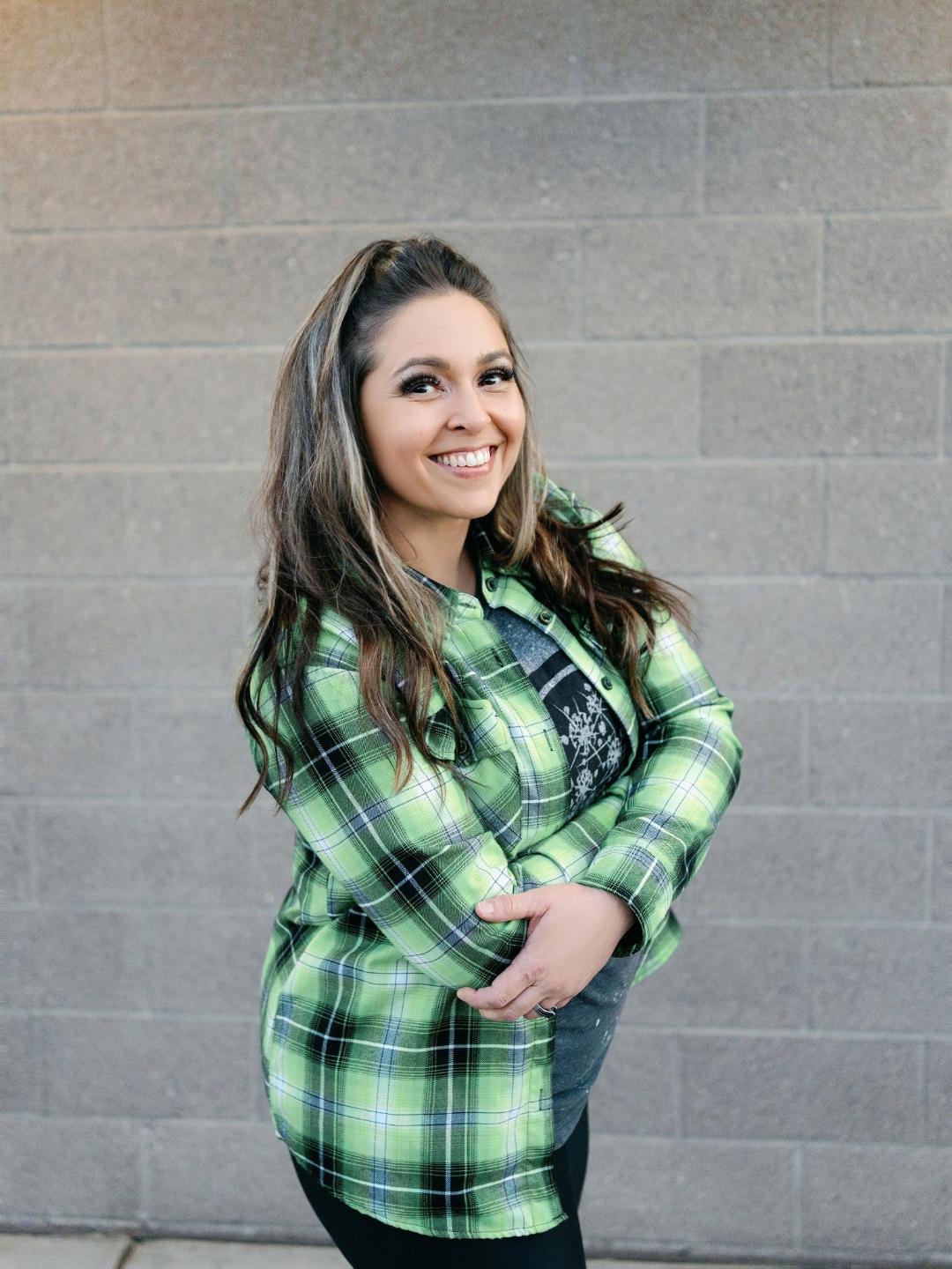 Smiling woman with long hair wearing a green plaid shirt standing against a gray brick wall.