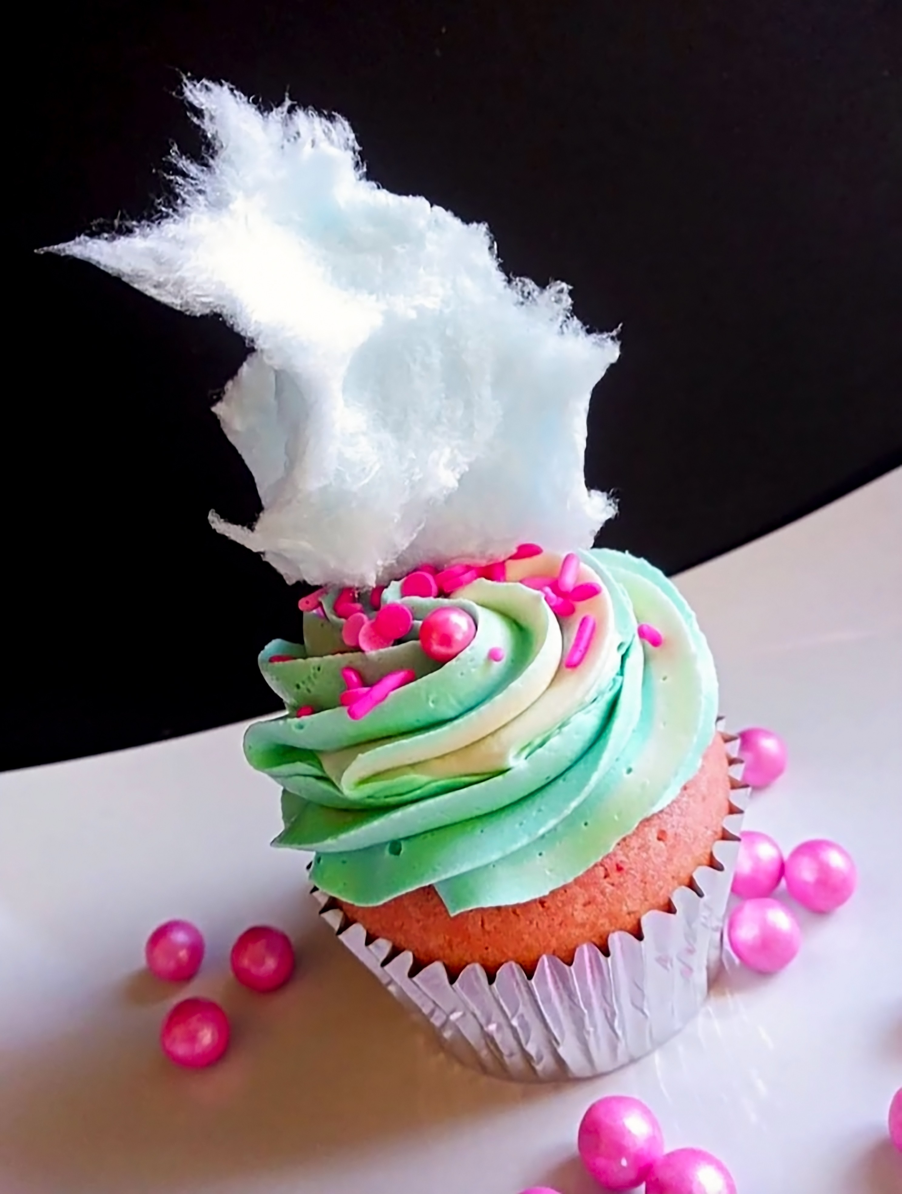 Cupcake with colorful swirled frosting and pink decorations, topped with cotton candy, on a white surface with pink pearls.