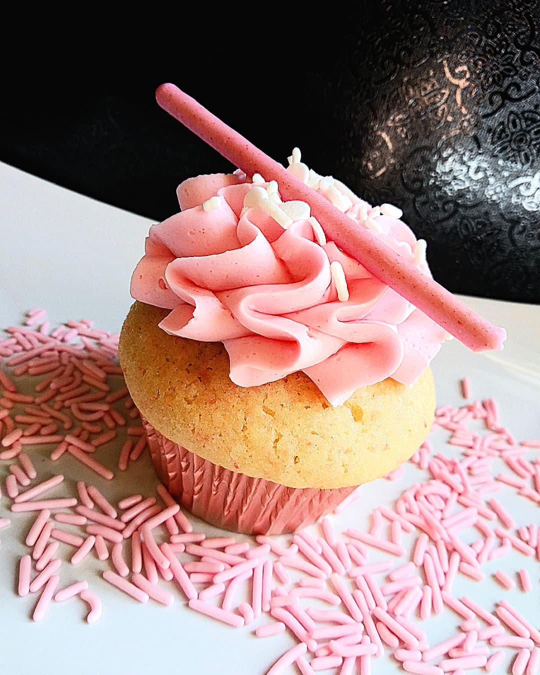 Decorated cupcake with pink frosting, a pink stick, and pink sprinkles on a white surface.
